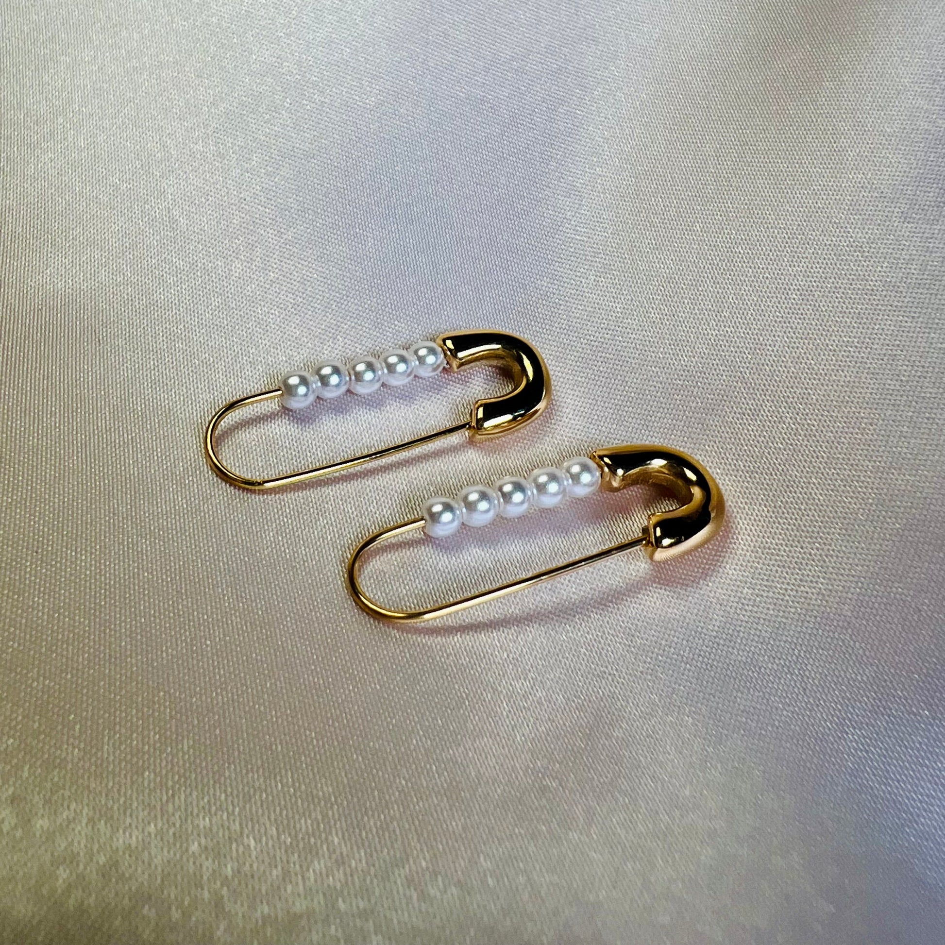 Gold safety pin earrings with pearls on a light fabric background